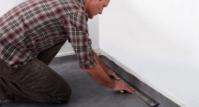 Man in checkered shirt uses long metal tool on the corners of a black linoleum floor.