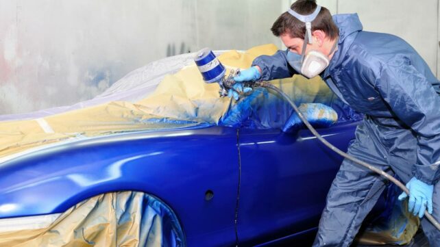 A technician, wearing protective gear, spraying paint on the body of a blue car in a workshop. - car respraying cost