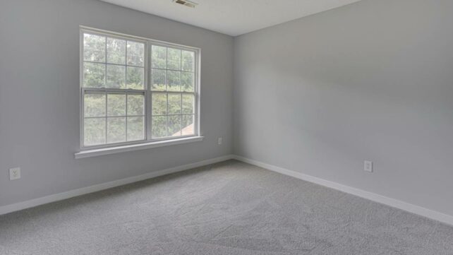 A spacious empty room featuring a large window that allows abundant natural light, with carpet flooring. - carpet laying cost