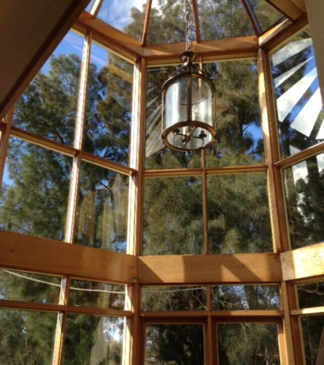 A tall, elegant glass window structure framed with natural wood, featuring a central hanging lantern and reflecting the surrounding trees and blue sky. - cost of window cleaning